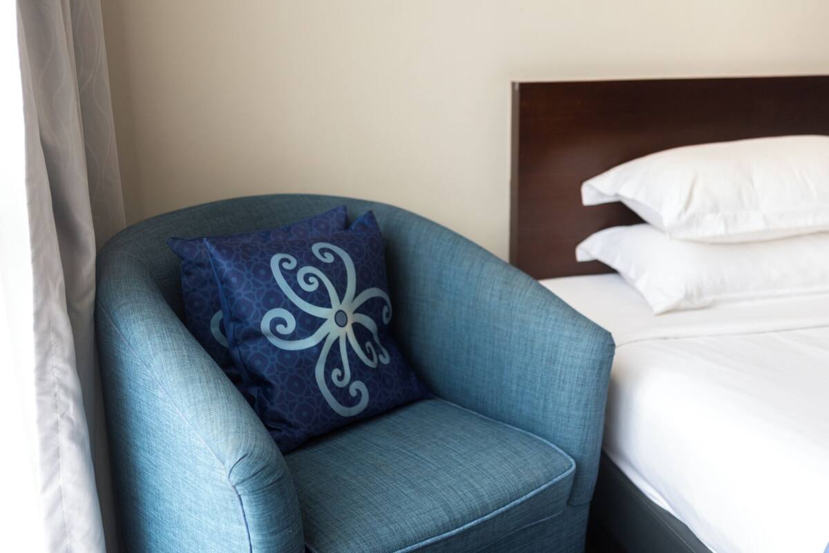 Una habitación de hotel con un sillón azul, una cama con sábanas blancas y un cabecero oscuro.