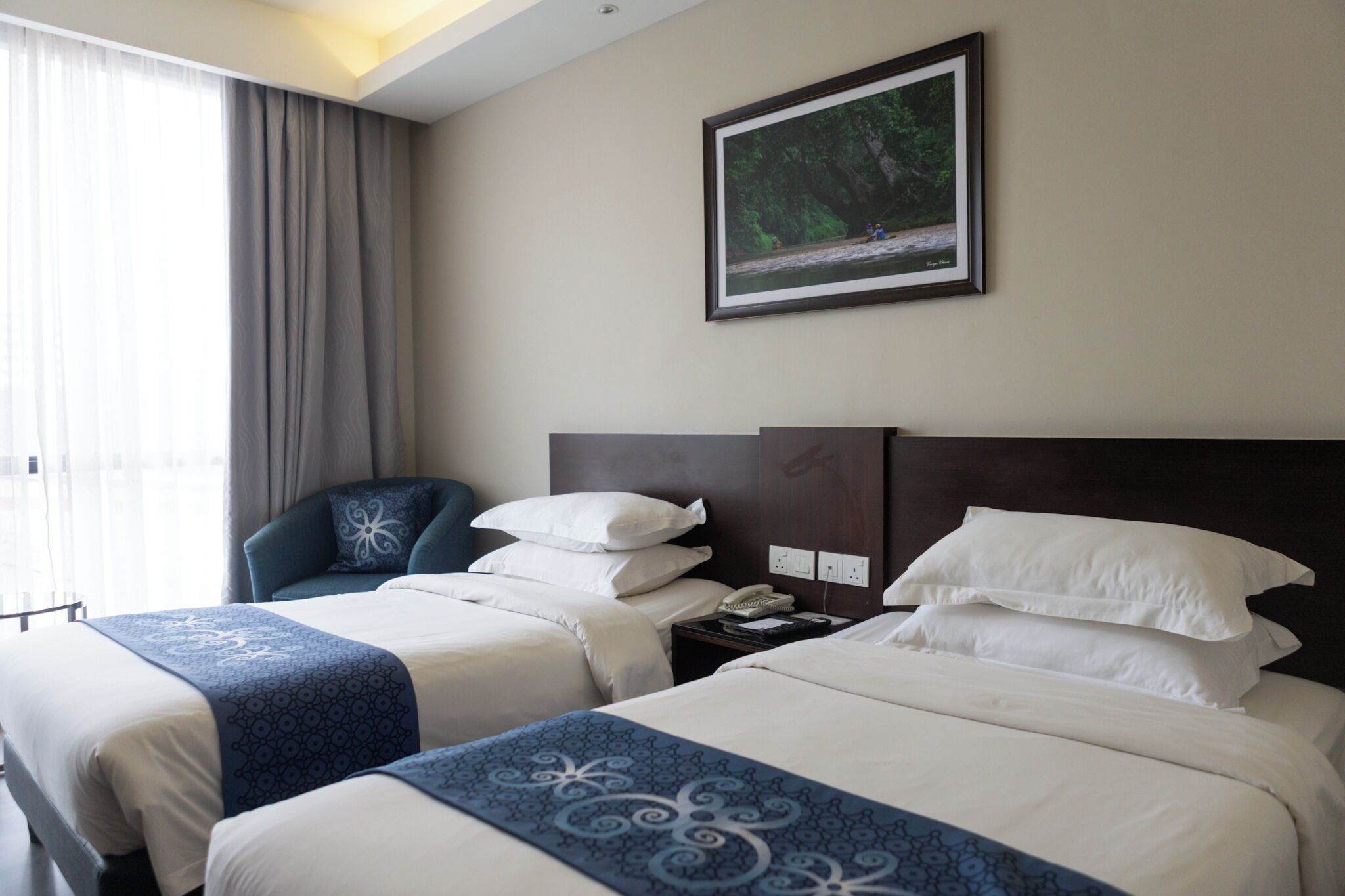 A hotel room with two beds, a desk, a chair, a painting, and a window with curtains.