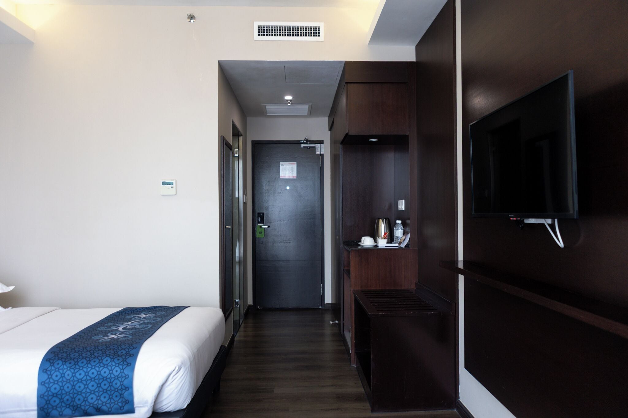 A hotel room with a bed, a TV mounted on the wall, a small table with a kettle, and a door leading to another room.