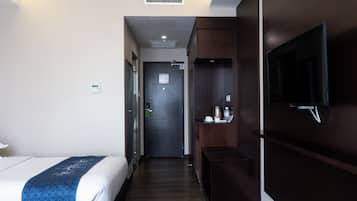 A hotel room with a bed, a TV mounted on the wall, a small table with a kettle, and a door leading to another room.