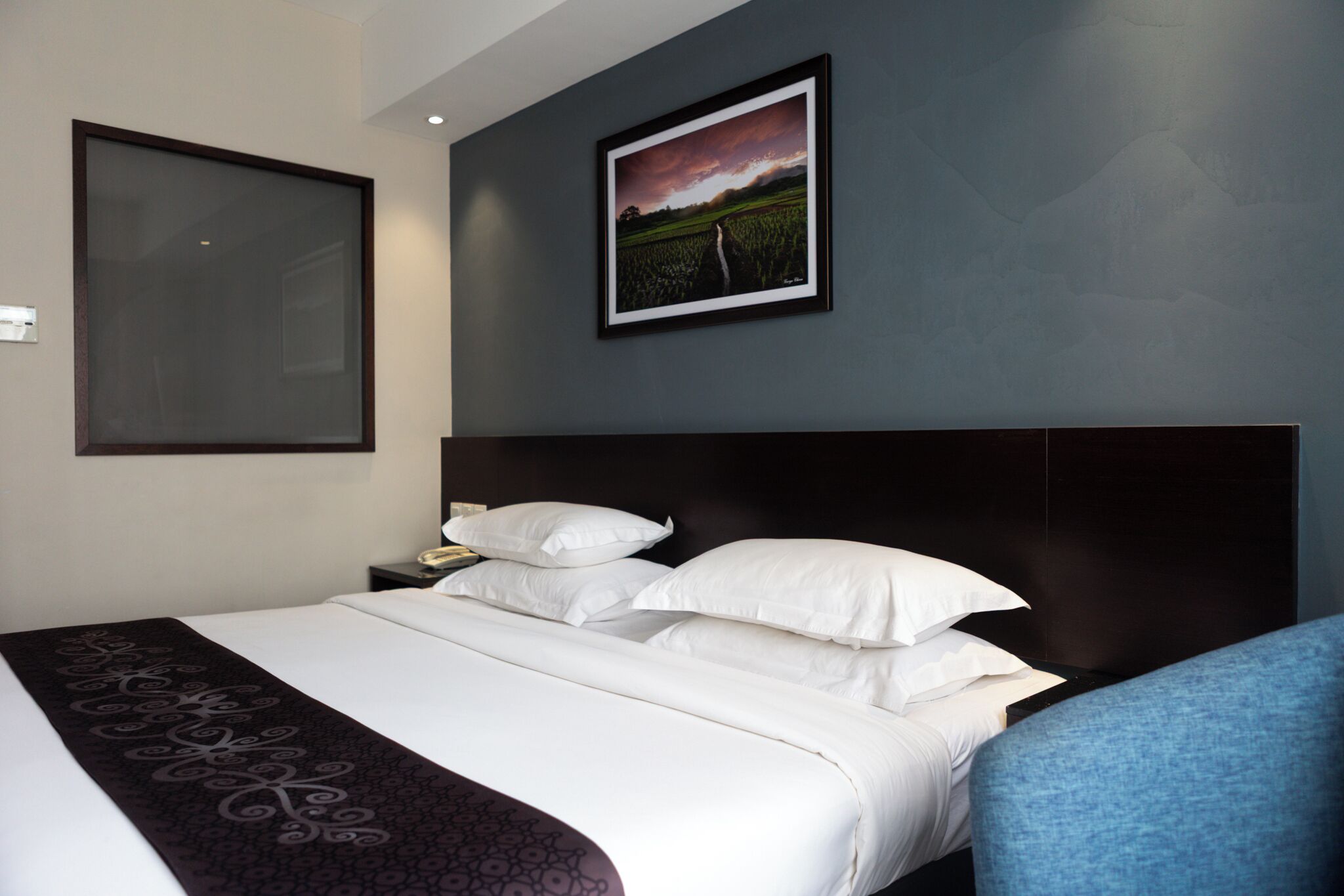 Habitación de hotel con una cama grande, cabecero, mesita de noche, un cuadro enmarcado en la pared y una silla azul.
