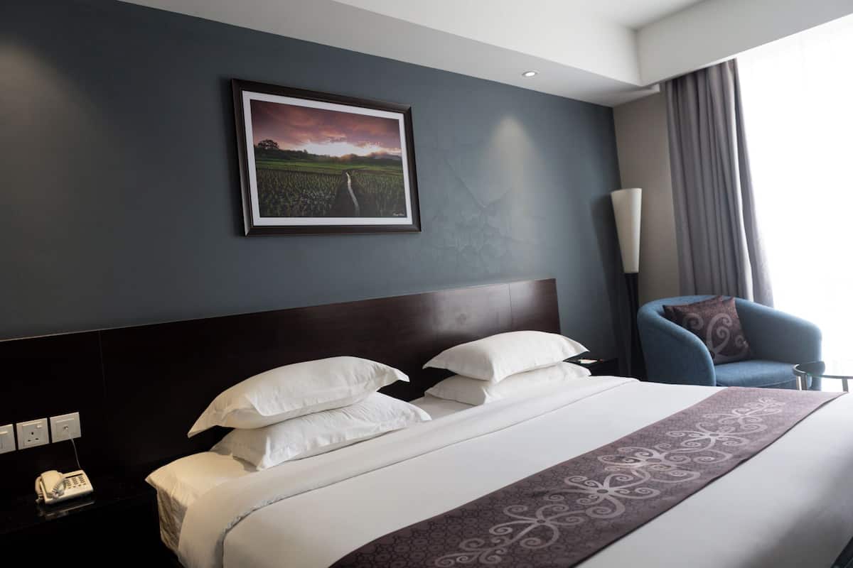 Una habitación de hotel con una cama grande, un cabecero, un cuadro enmarcado en la pared, un sillón azul y una mesita de noche con una lámpara.