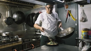 A chef in a professional kitchen with stainless steel surfaces, pots, and a wok. - Meritin Hotel (Kuching)