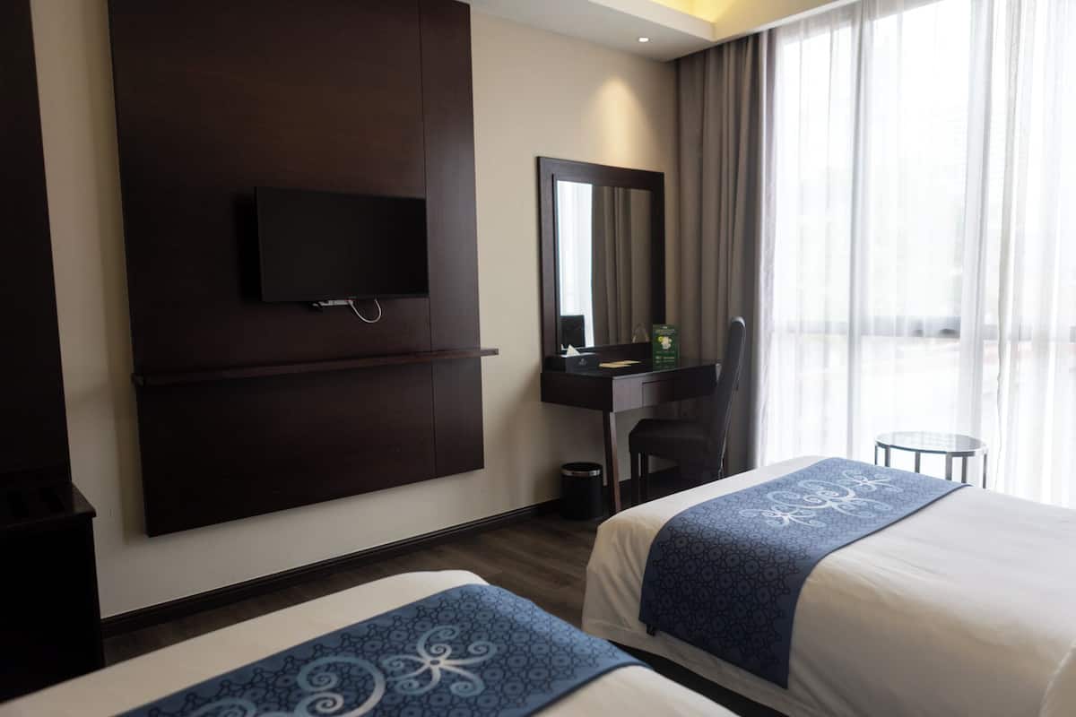 Habitación de hotel con dos camas, un escritorio, una silla, un televisor montado en la pared y un ventanal con cortinas.