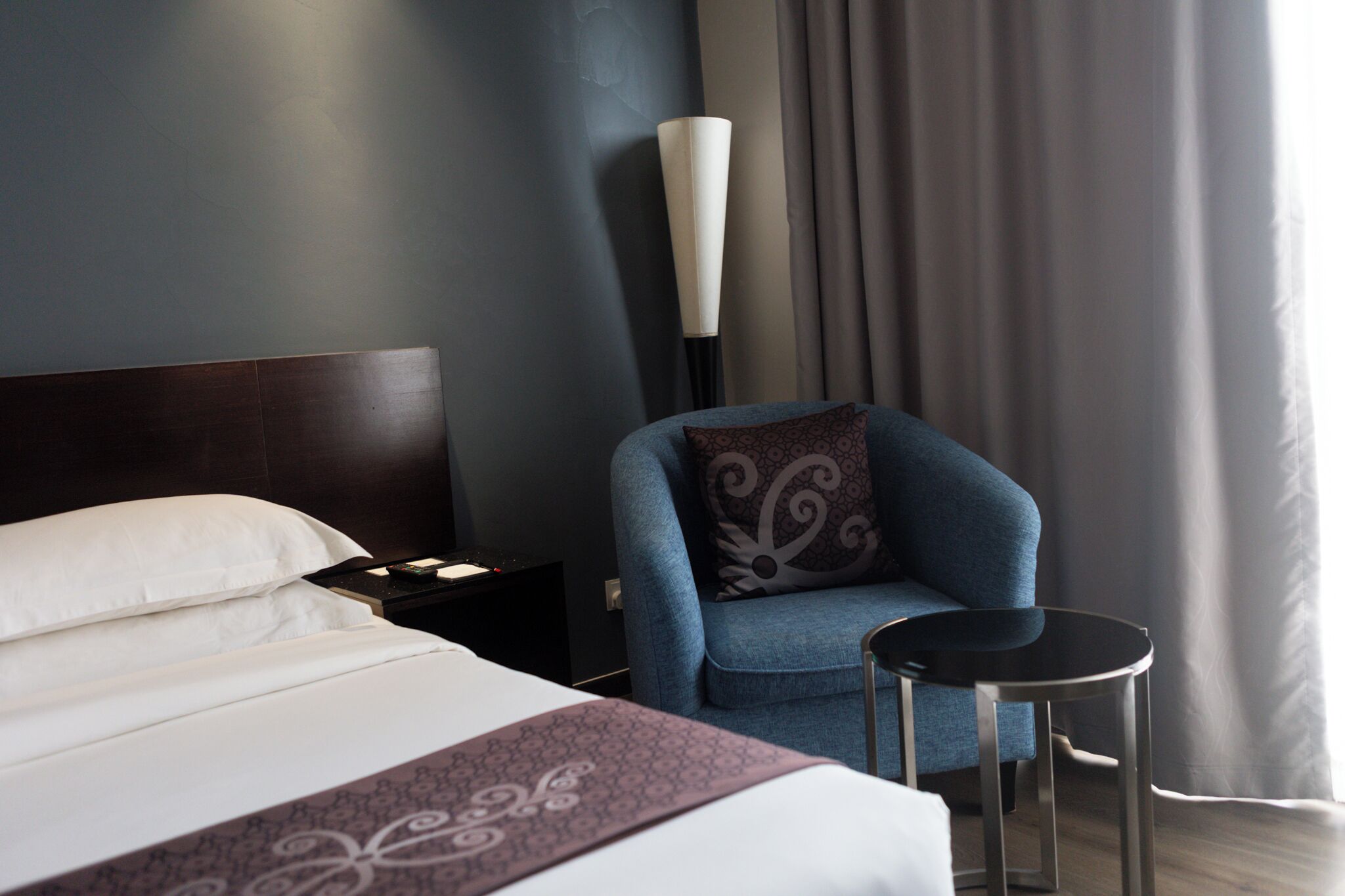 A hotel room with a bed, a blue armchair, a small round table, and a tall floor lamp.