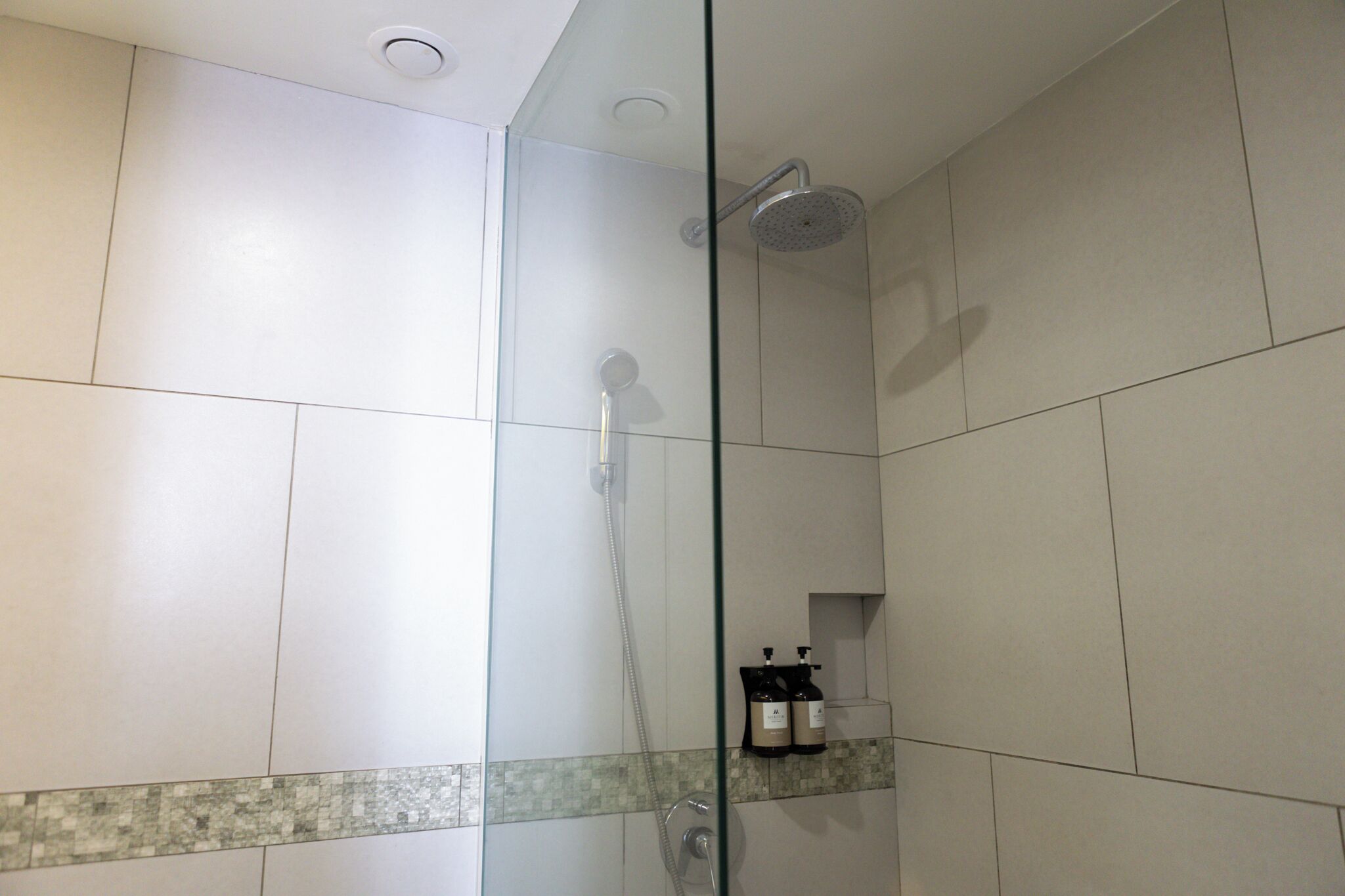 Een douche met glazen wand, een handdouche, een muurkraan en een zeepdispenser.