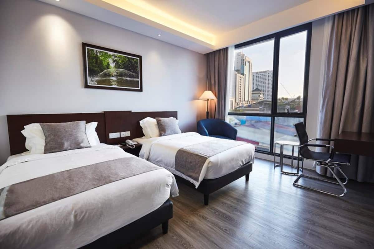 A hotel room with two beds, a desk, a chair, a lamp, and a large window with a city view.