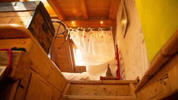 Uma escada de madeira leva a um quarto com uma cama, uma janela com cortinas e um pequeno objeto decorativo na parede.