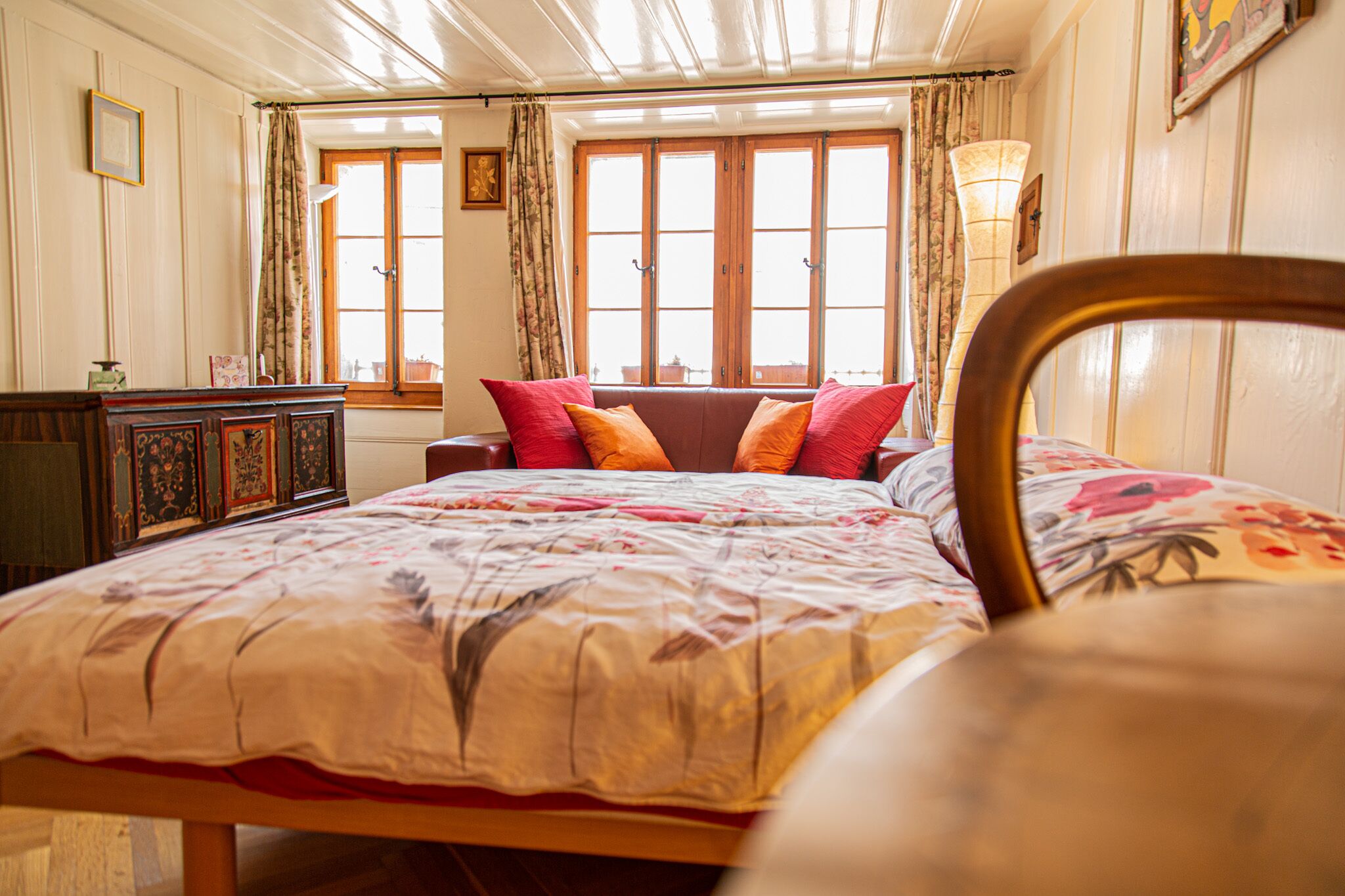 Ein Schlafzimmer mit einem Bett, einem hölzernen Kopfteil, einer Kommode und einem Fenster mit Vorhängen.