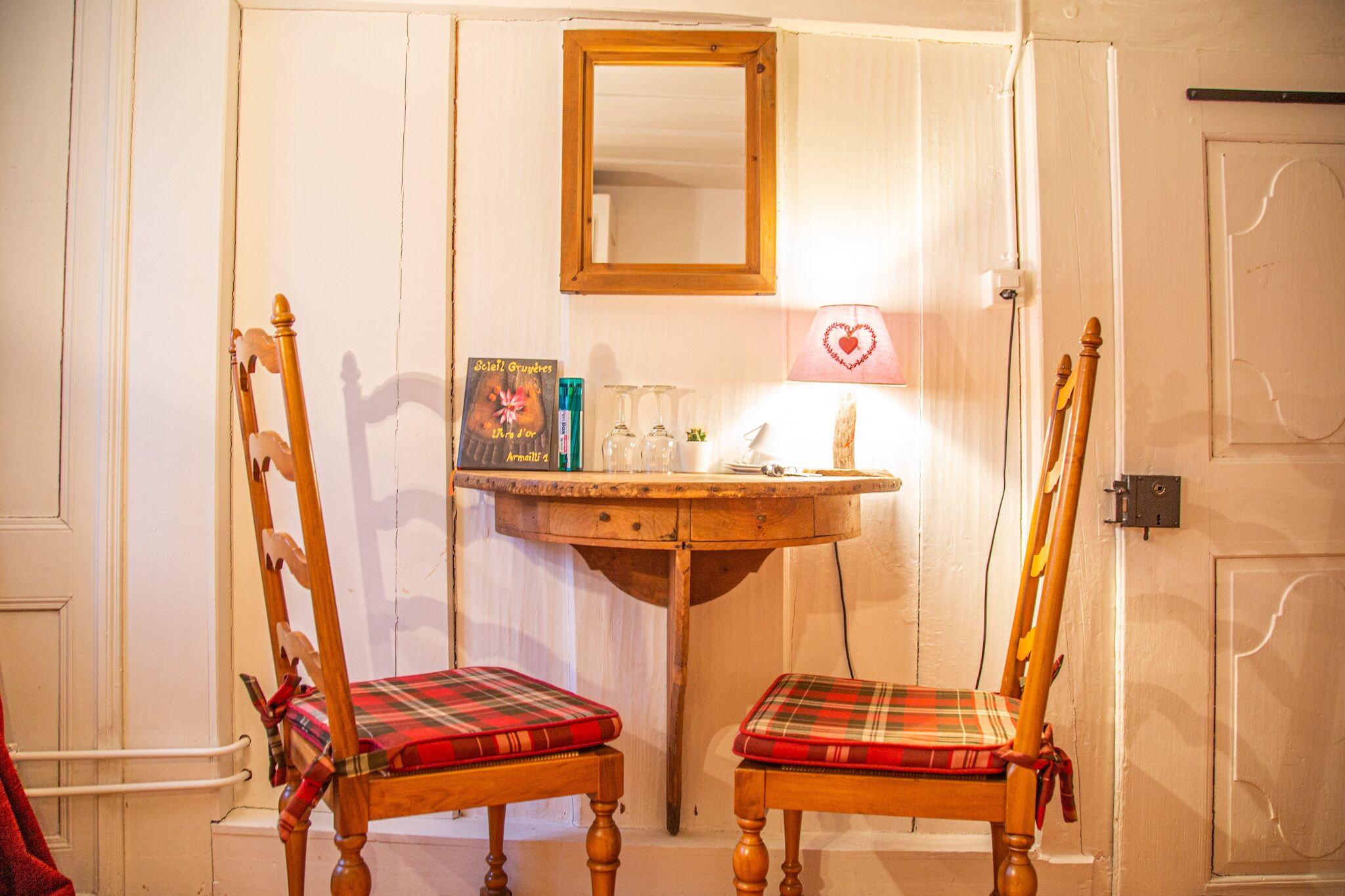 Una habitación con dos sillas de madera con cojines escoceses, una pequeña mesa de madera y un espejo sobre ella, además de una lámpara de pared con forma de corazón.