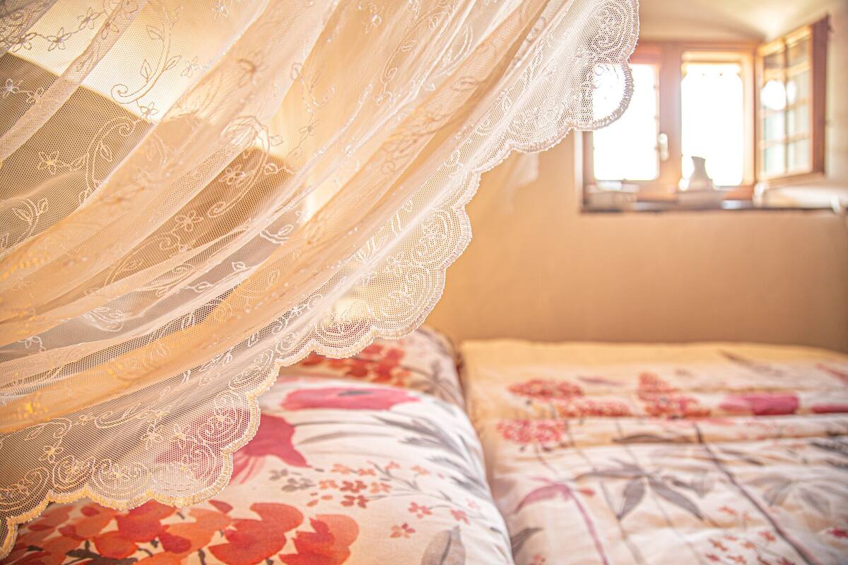 Ein Schlafzimmer mit einem blumengedeckten Bett, einem Spitzenbaldachin und einem Fenster mit Sonnenschein.