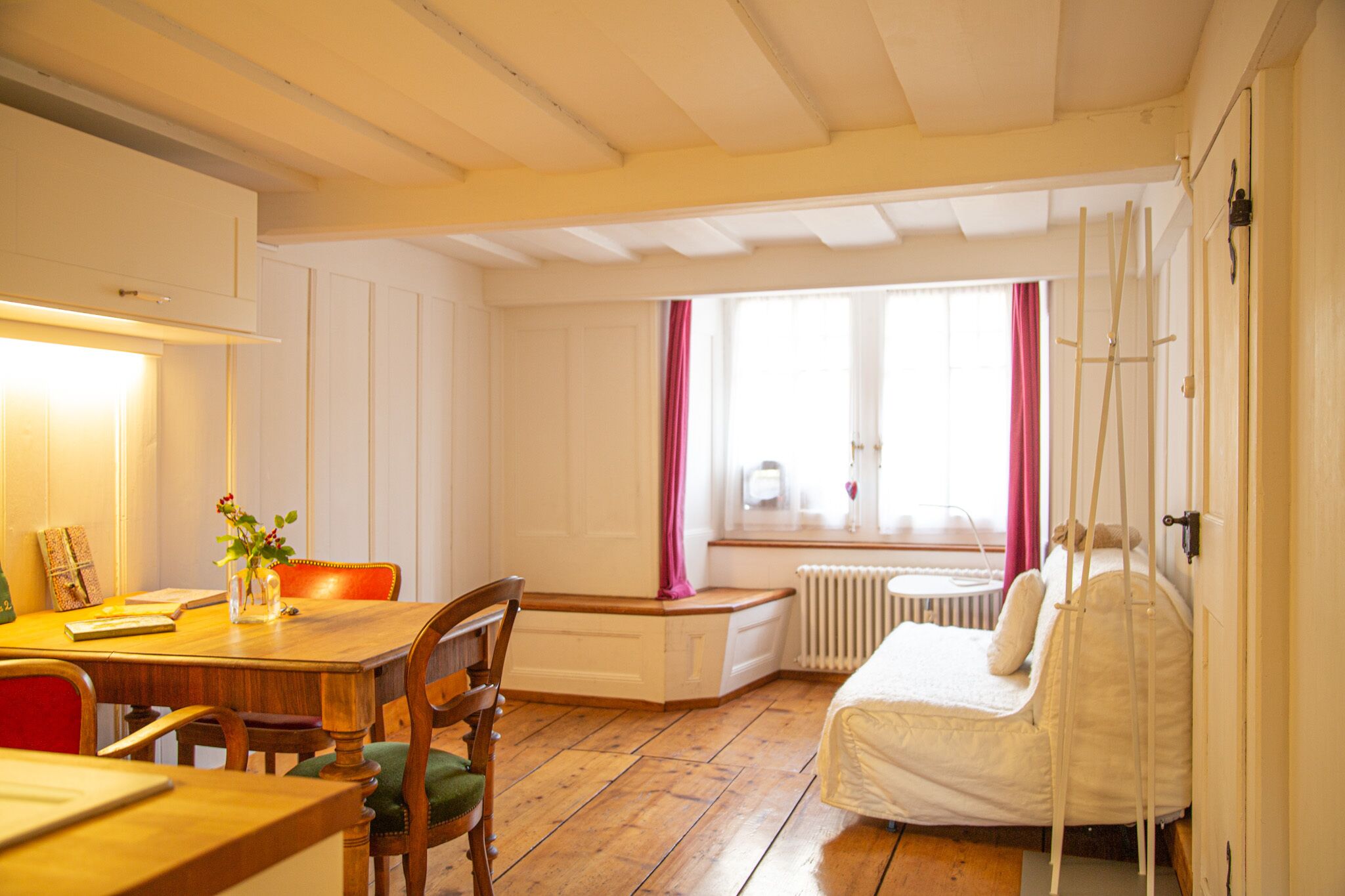 Une pièce comprenant une table à manger en bois, des chaises, un lit avec un matelas blanc, une fenêtre avec des rideaux roses et un radiateur.