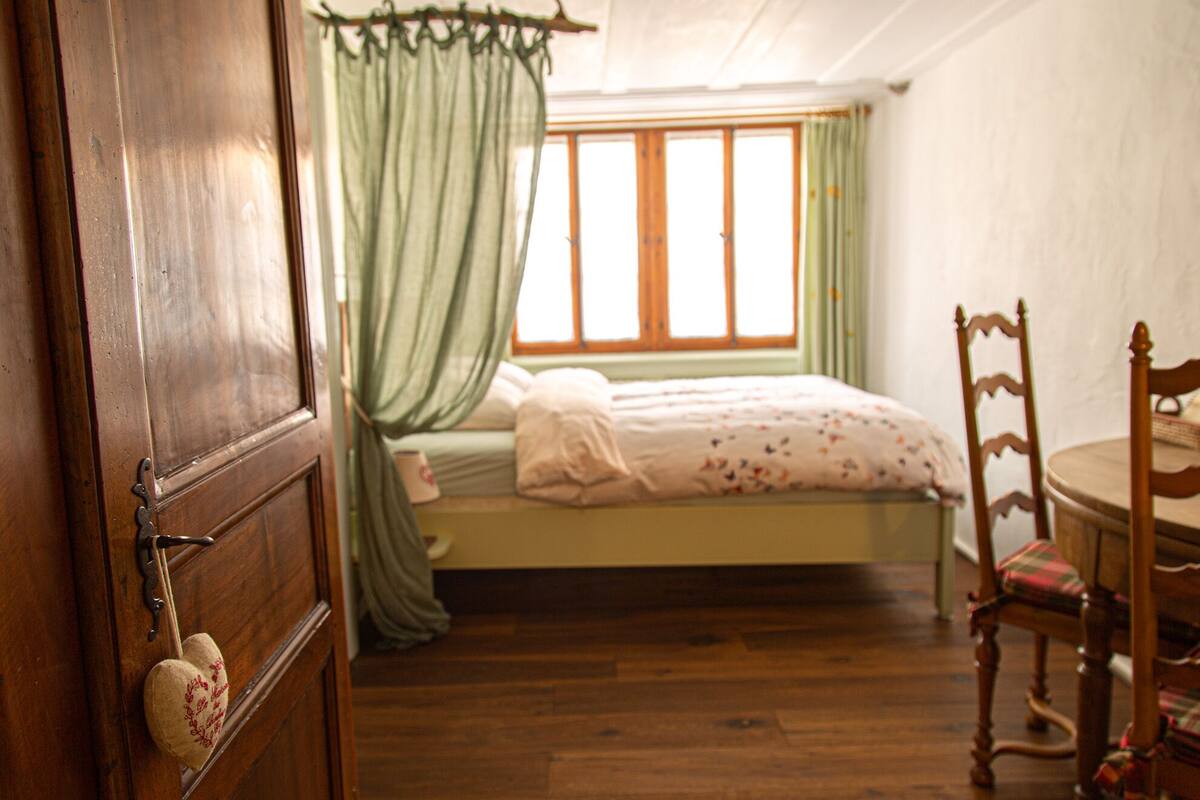 Ein Schlafzimmer mit einem hölzernen Bett, einem grünen Baldachin, einem Holzstuhl und einem Fenster mit Vorhängen.