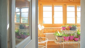 Una cabaña de madera con vistas a las montañas a través de las ventanas. Hay varias macetas con plantas sobre una mesa y un radiador al fondo.