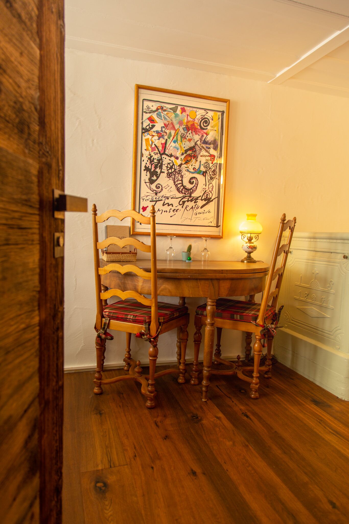 Uma área de refeições com mesa e cadeiras de madeira, uma obra de arte emoldurada acima da mesa e um abajur aceso.