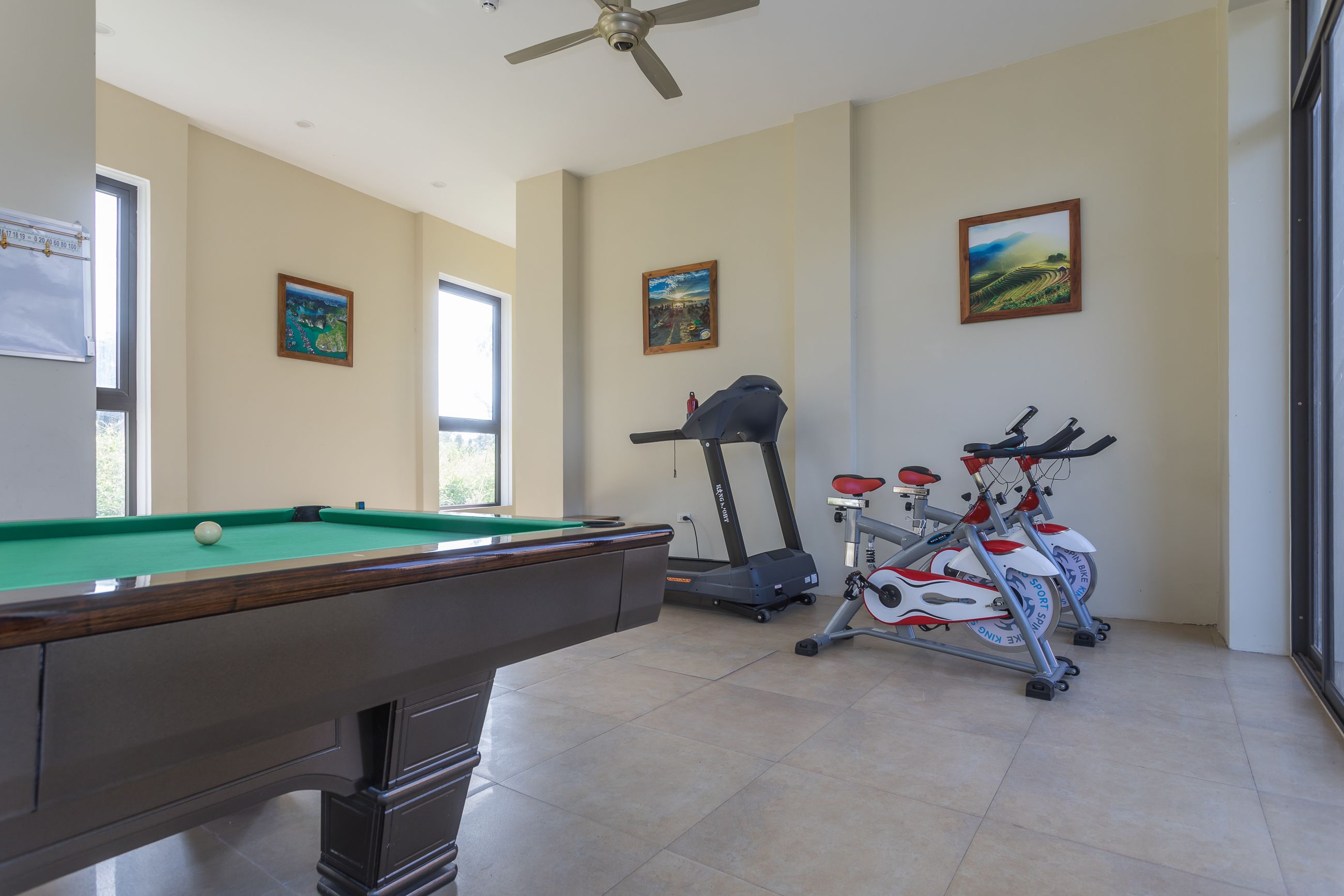 A pool table, exercise bikes, and a ceiling fan in a room with large windows and artwork on the walls.