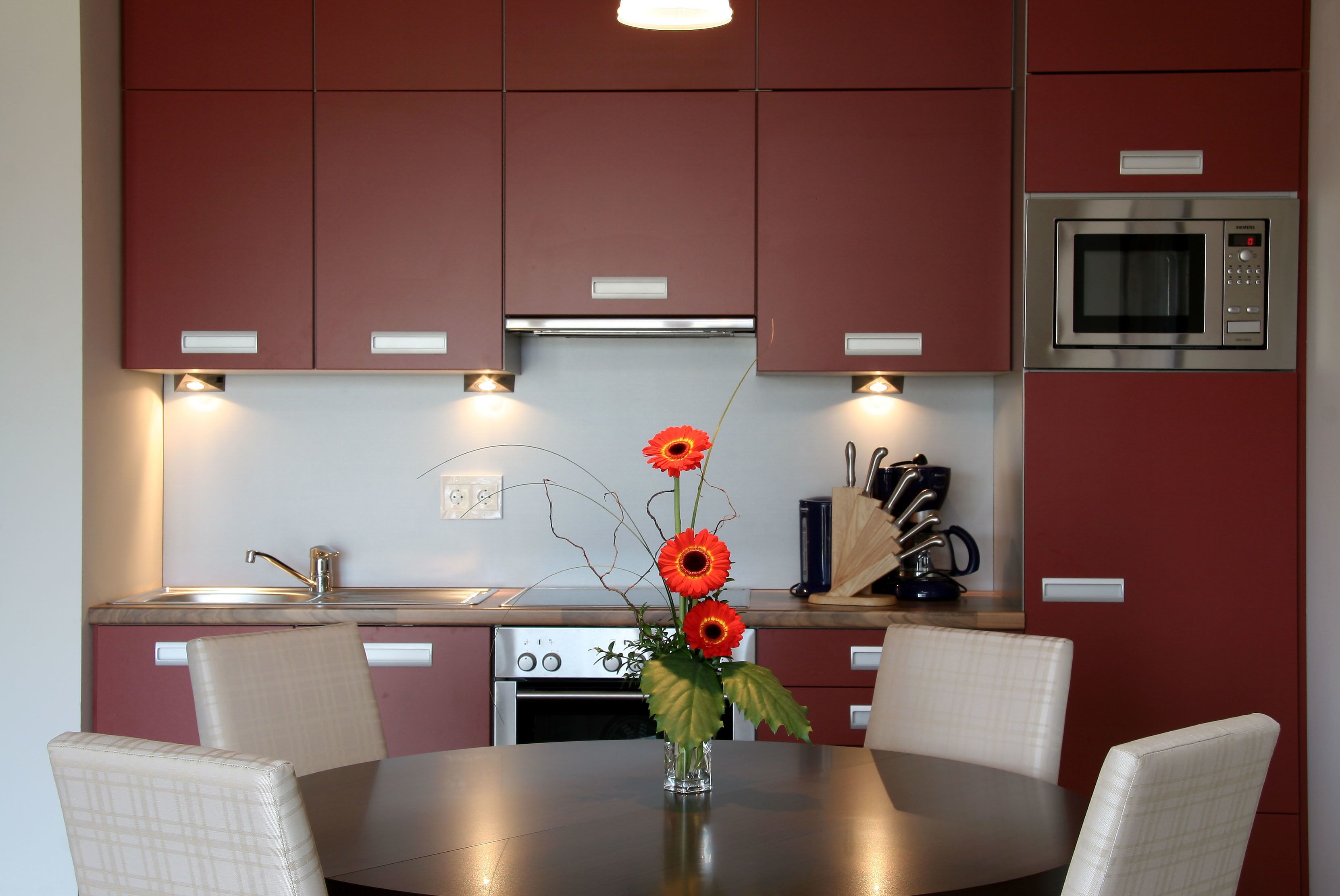 A modern kitchen with red cabinets, a stainless steel microwave, and a dining table with four chairs.