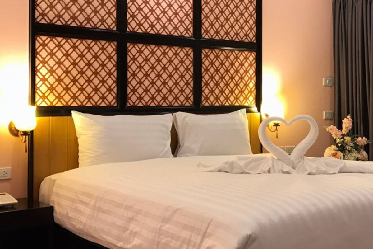 A hotel room with a bed, two white pillows, a decorative heart-shaped swan on the bed, a nightstand with a phone, and a wall with a lattice design.