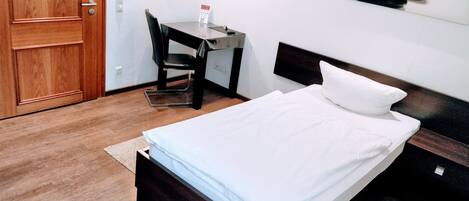 A hotel room with a wooden door, a desk with a chair, a bed with white linens, and a wall-mounted artwork.
