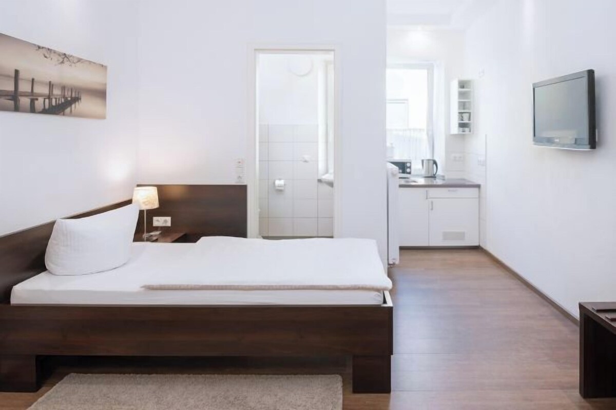 Een slaapkamer met een bed, een bedlampje, een televisie en uitzicht op een badkamer en keuken.