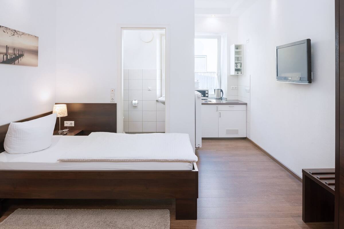 Une chambre d’hôtel avec un lit en bois, une lampe de chevet, un téléviseur à écran plat et une vue sur la salle de bain et la cuisine.