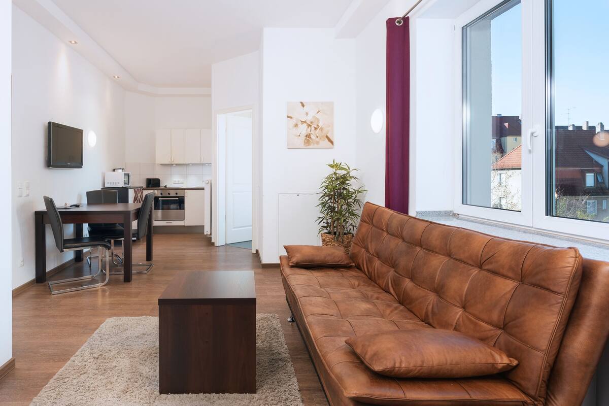 Un salon moderne avec un canapé en cuir marron, une table à manger avec des chaises, un coin cuisine et une grande fenêtre donnant sur des immeubles.