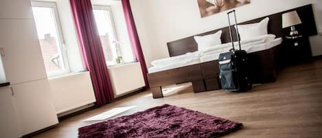 A hotel room with a bed, a suitcase, a window with curtains, a radiator, and a painting on the wall.