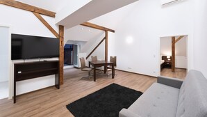 Ein modernes Wohnzimmer mit Fernseher, Holzbalken, einem Esstisch mit Stühlen und einem grauen Sofa.