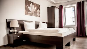 Ein modernes Schlafzimmer mit einem großen Bett, einem dunklen Holz-Kopfteil, einem Nachttisch mit Lampe, einem Kleiderschrank und einem Fenster mit Vorhängen.