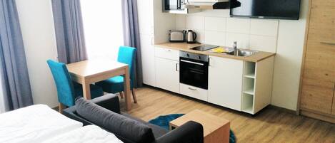 A compact hotel room with a bed, a small kitchenette, a dining table with chairs, and a TV mounted on the wall.