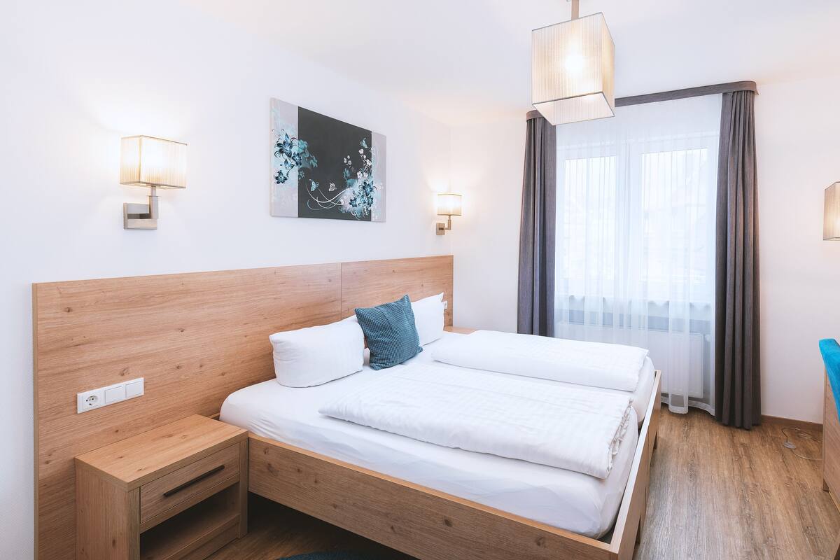 A hotel room with a wooden bed, a desk, and a chair. There is a window with curtains, a wall-mounted TV, and a painting on the wall.