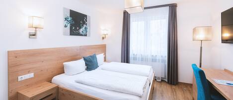 Habitación de hotel con cama de madera, escritorio y silla. Cuenta con una ventana con cortinas, un televisor montado en la pared y un cuadro.