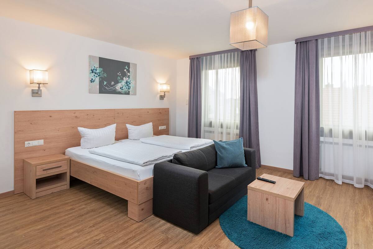 A modern hotel room with a wooden headboard bed, a black sofa, a small wooden side table, and a desk with a chair.