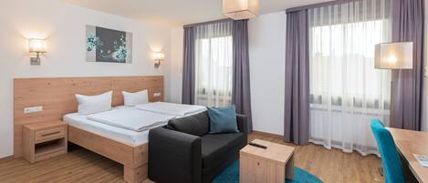 A modern hotel room with a wooden headboard bed, a black sofa, a small wooden side table, and a desk with a chair.