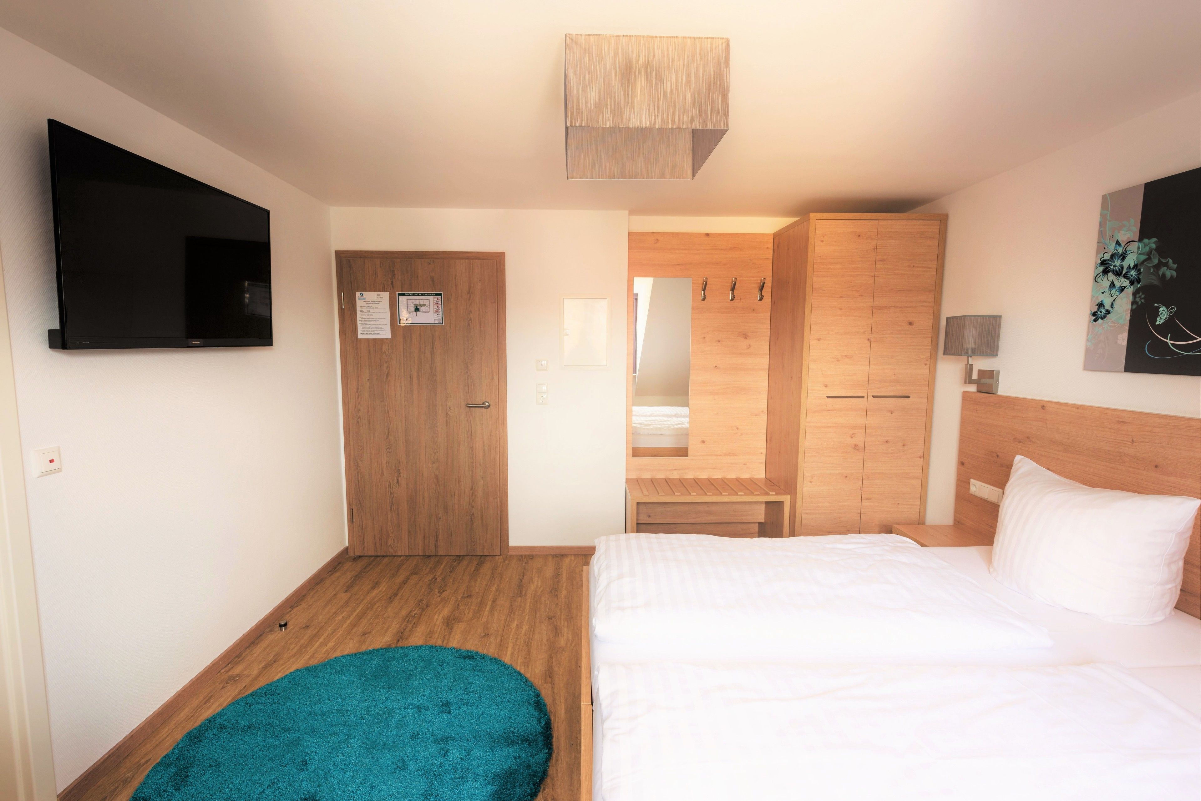 A bedroom with a bed, a TV mounted on the wall, a wooden wardrobe, and a small table with a lamp.