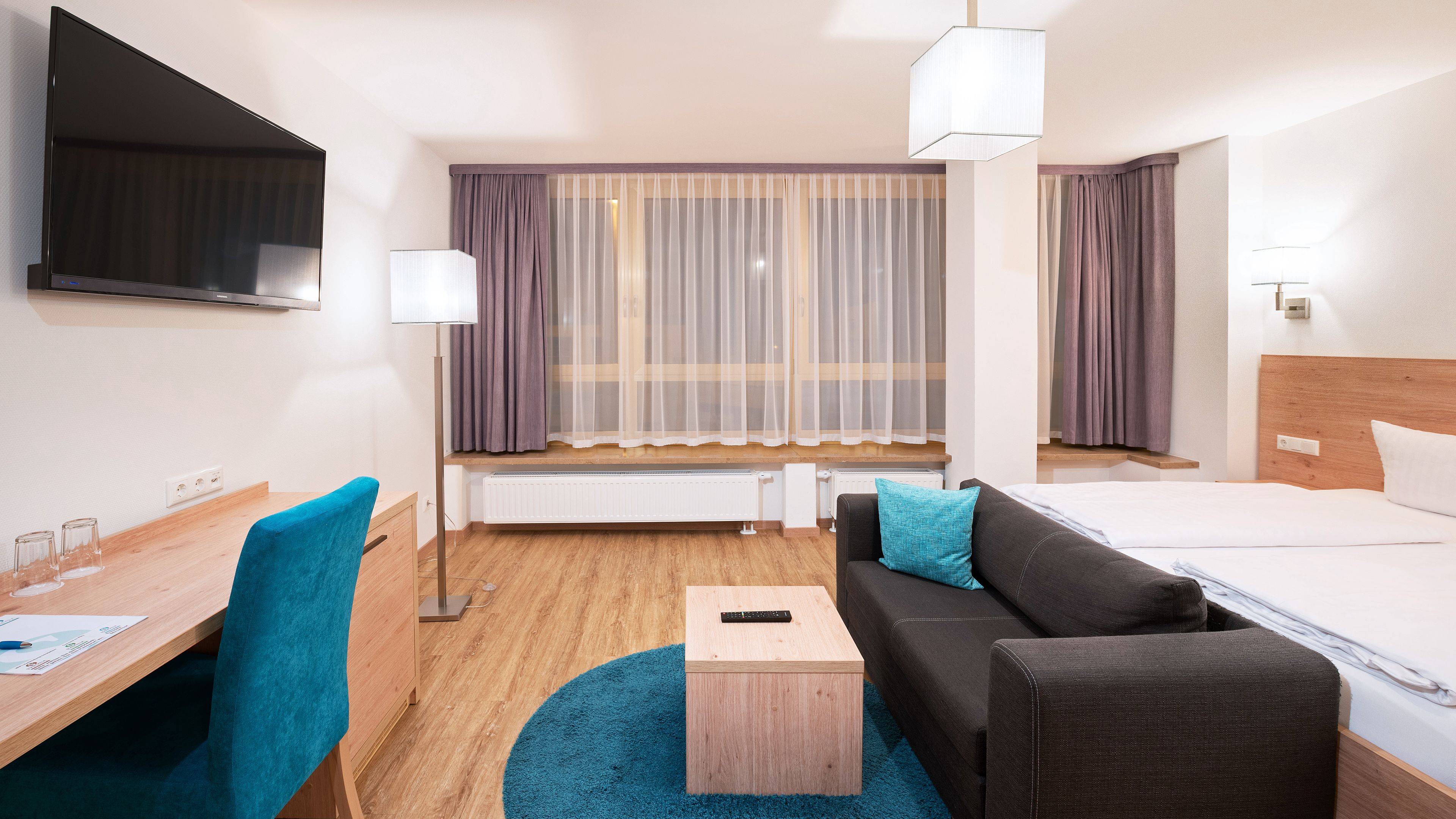 A hotel room with a bed, a desk with a chair, a small table, a sofa, a TV, and a window with curtains.