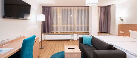 Una habitación de hotel con cama, escritorio con silla, una mesita, sofá, televisor y ventana con cortinas.