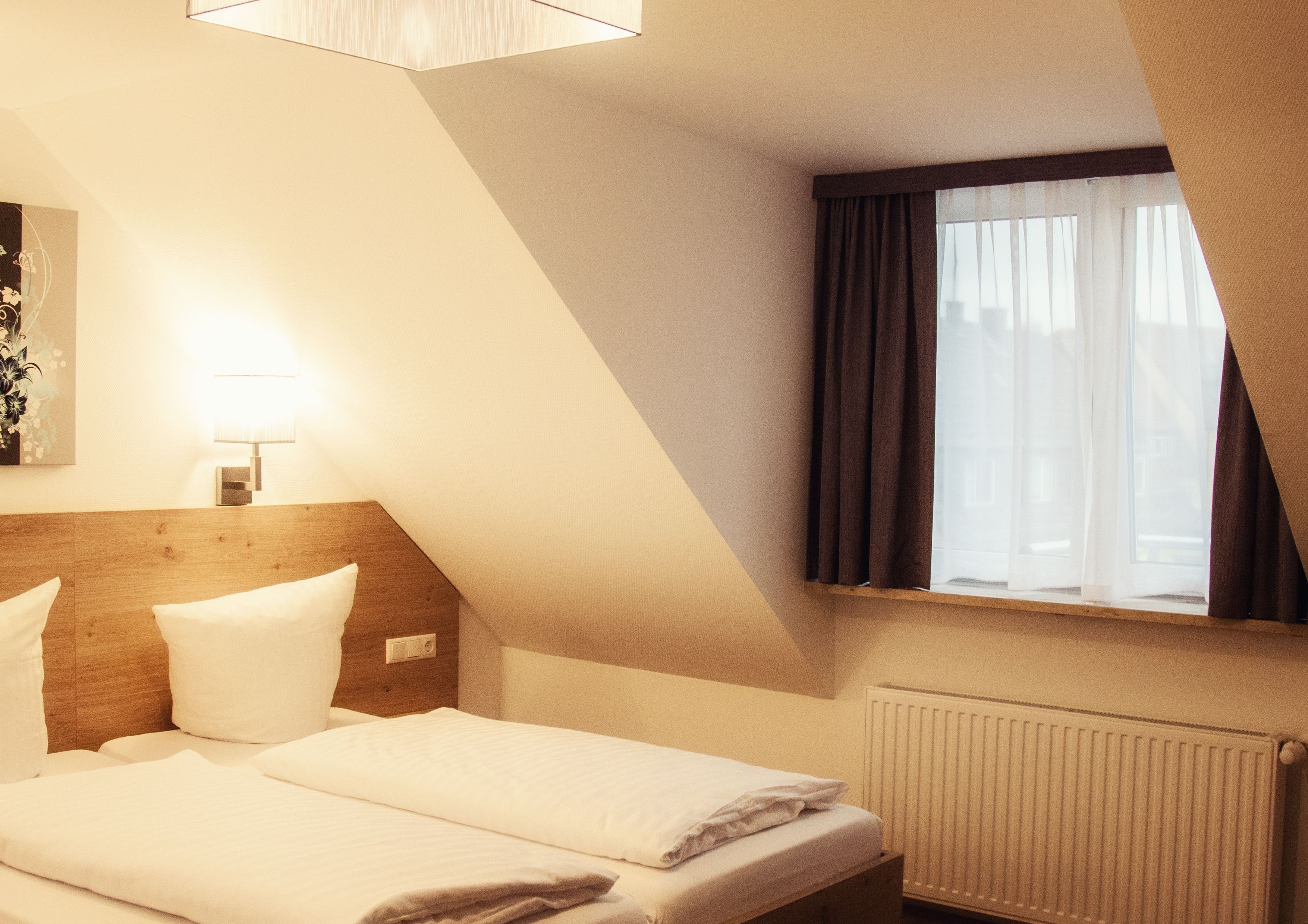 A hotel room with a bed, a nightstand, a lamp, a window with curtains, and a radiator.