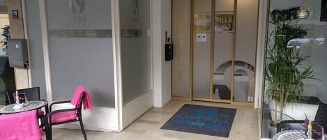 Un hall de hotel con mostrador de recepción, zona de estar con sillas y una mesa, puerta de cristal y felpudo con el nombre del hotel.