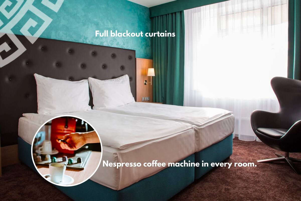 A hotel room with full blackout curtains, a tufted headboard, a Nespresso coffee machine, and a modern chair.
