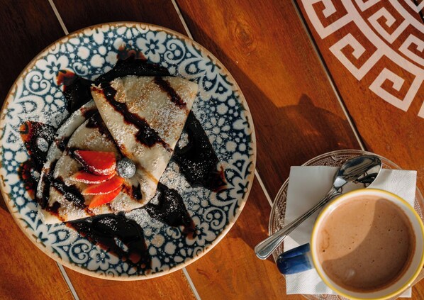 Un plato de comida con un postre, posiblemente una crepa o panqueque, cubierto de salsa de chocolate y fruta, acompañado de una taza de café sobre una mesa con mantel estampado.