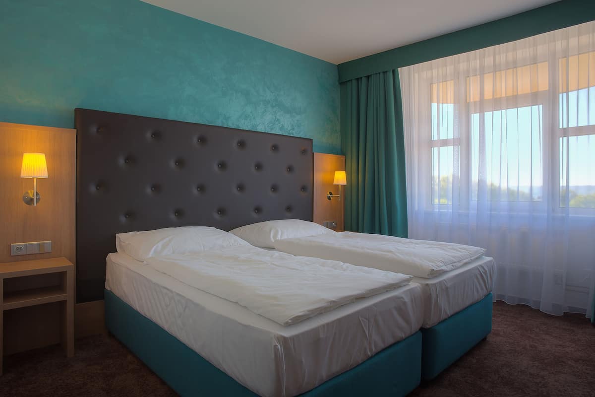 A hotel room with a large bed, two bedside tables, and a view of the outdoors through sheer curtains.