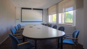 Un salón de conferencias con una gran mesa, sillas azules, una pantalla de proyector y ventanas con persianas.