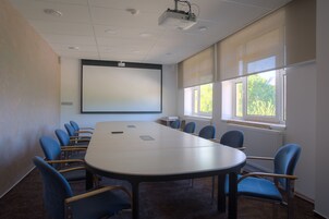 Un salón de conferencias con una gran mesa, sillas azules, una pantalla de proyector y ventanas con persianas.
