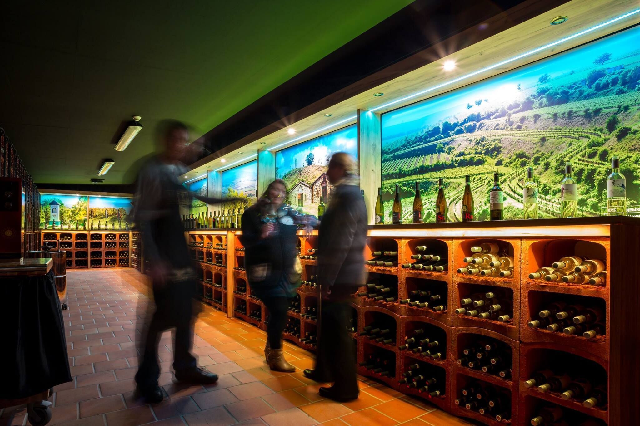 A wine cellar with wooden racks filled with bottles, a large mural of a vineyard, and a brick floor.