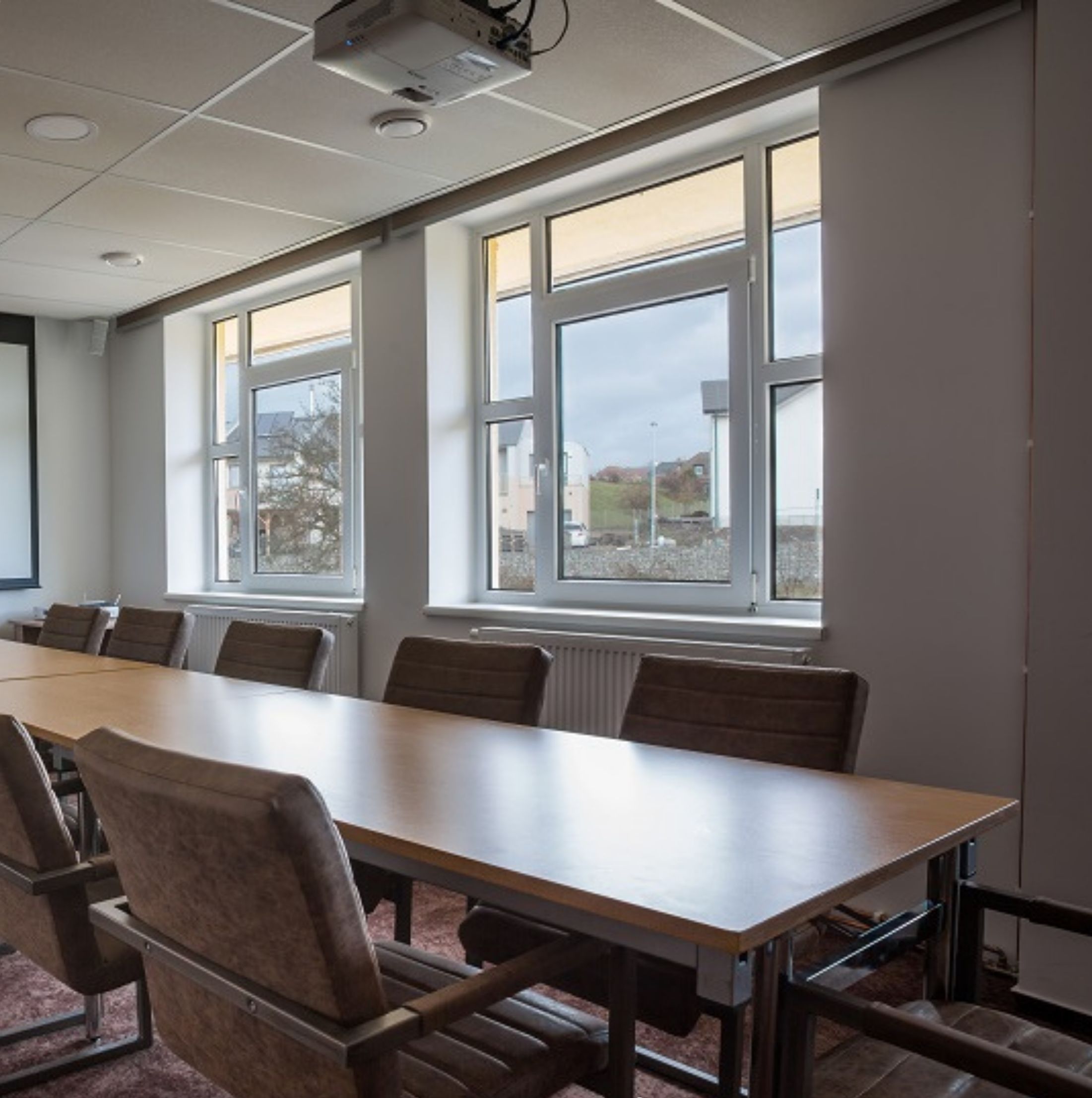 Un salón de conferencias con una larga mesa y sillas, ventanas amplias y una pantalla de proyector.