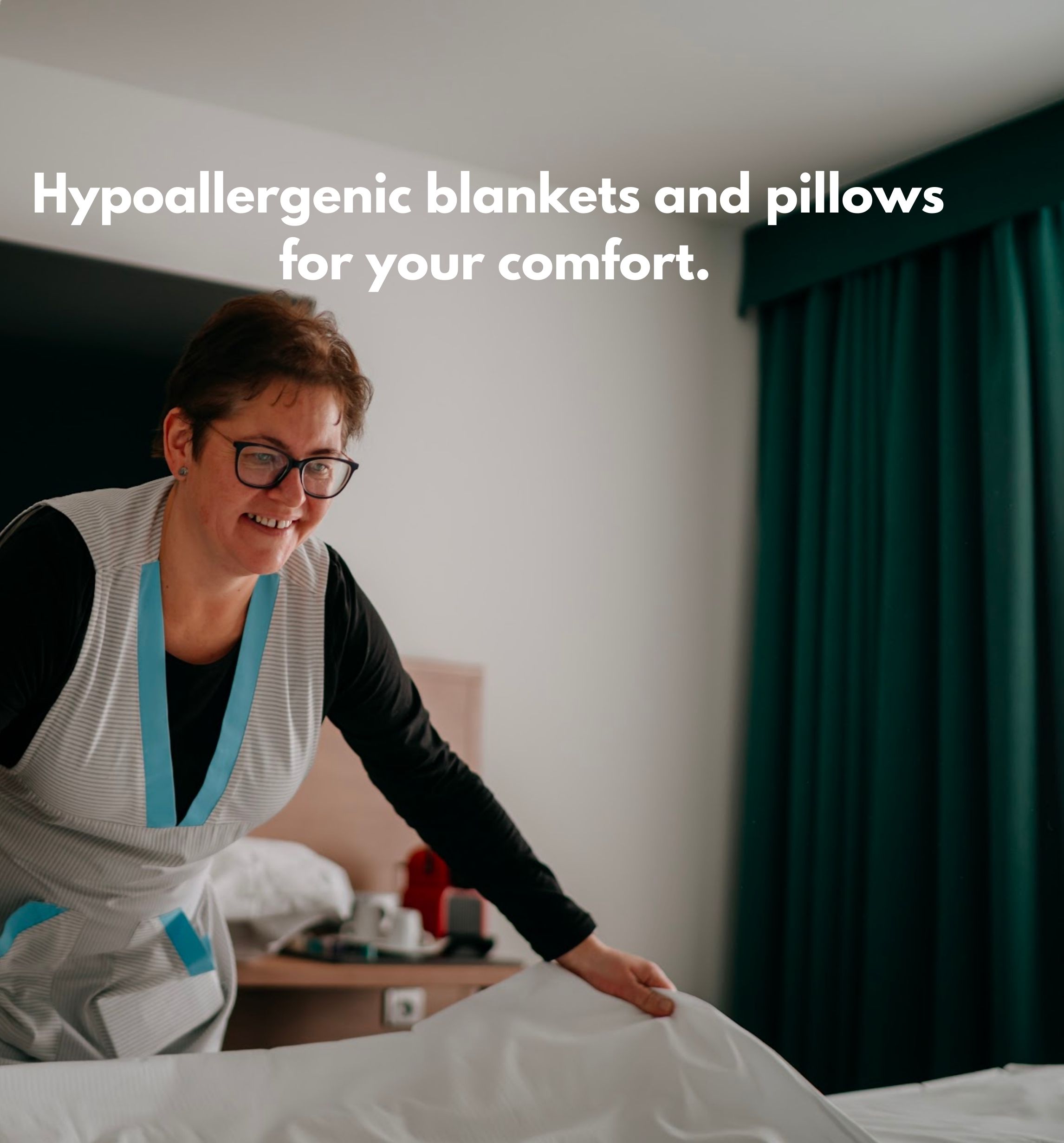 A person adjusting a bed in a hotel room with hypoallergenic blankets and pillows.