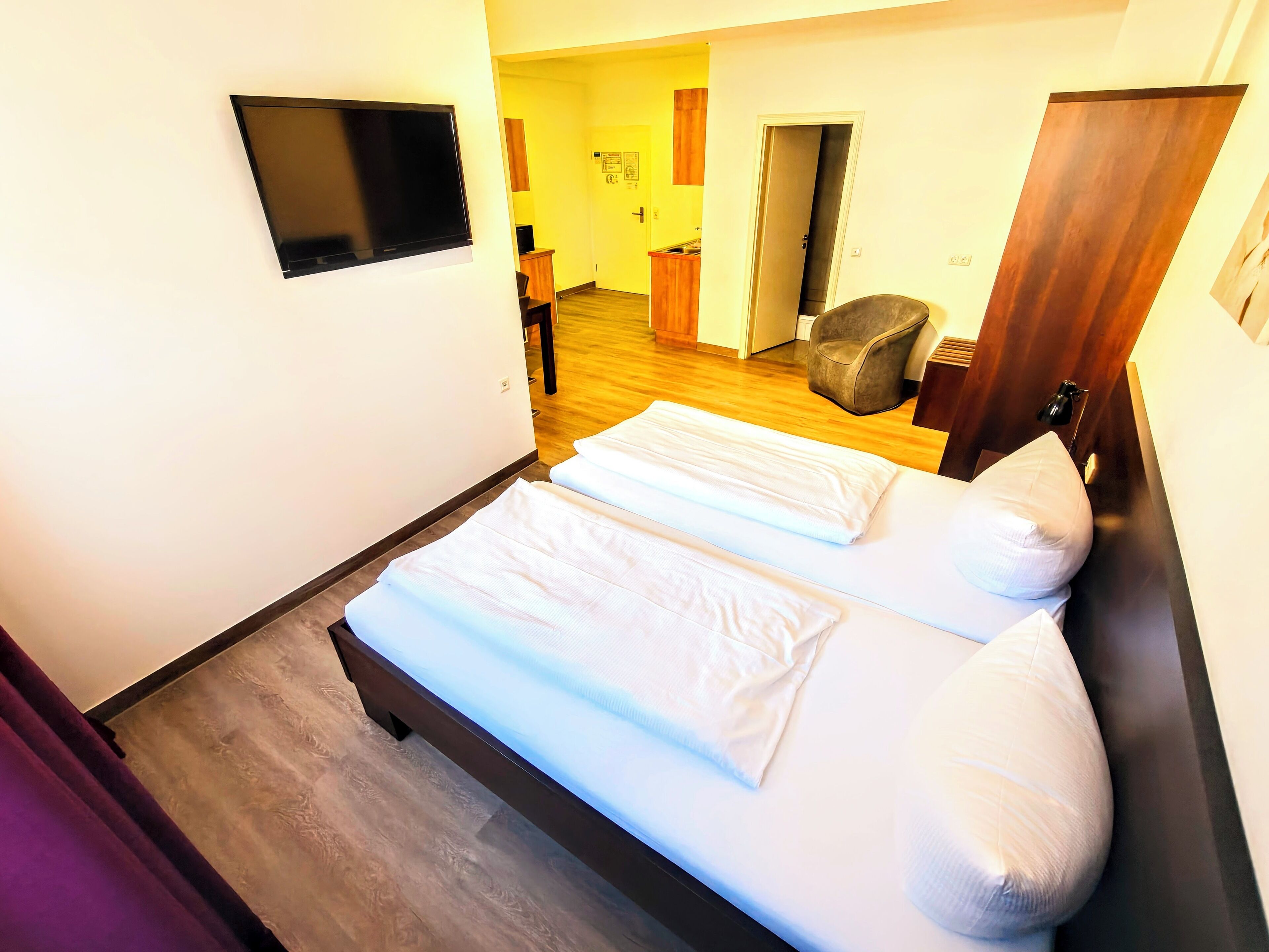 A hotel room with two beds, a TV mounted on the wall, a wooden floor, and a door leading to a bathroom.