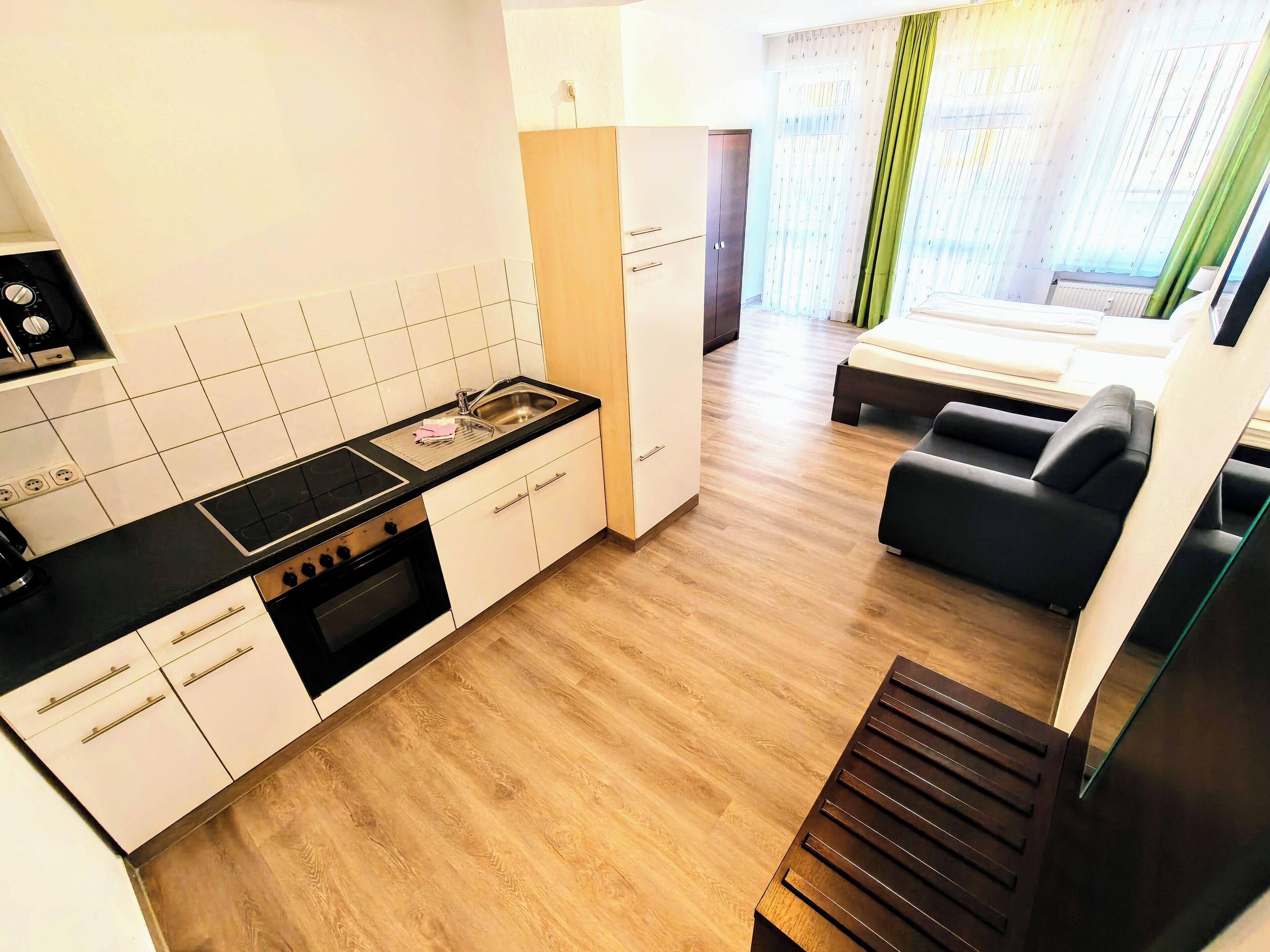 A modern kitchen with white cabinets, a black countertop, and a built-in oven. There is a dining area with a black sofa and a glass table. The room has wooden flooring and large windows with sheer curtains.