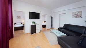 Une chambre d’hôtel moderne avec un canapé en cuir noir, un téléviseur à écran plat, un petit bureau et un lit avec une literie blanche.